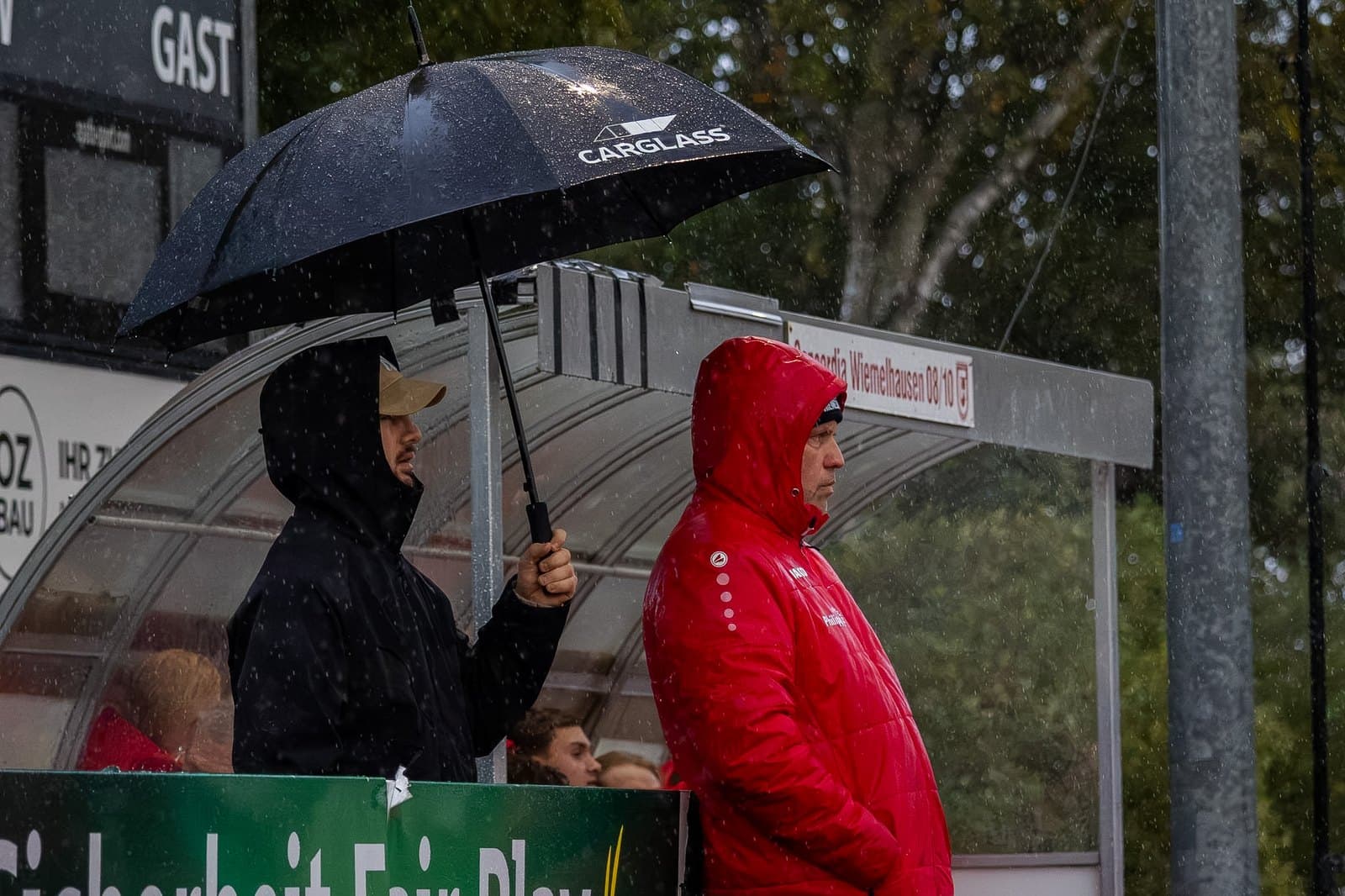 Zuschauer unter Regenschirmen am Spielfeldrand im Sportpark