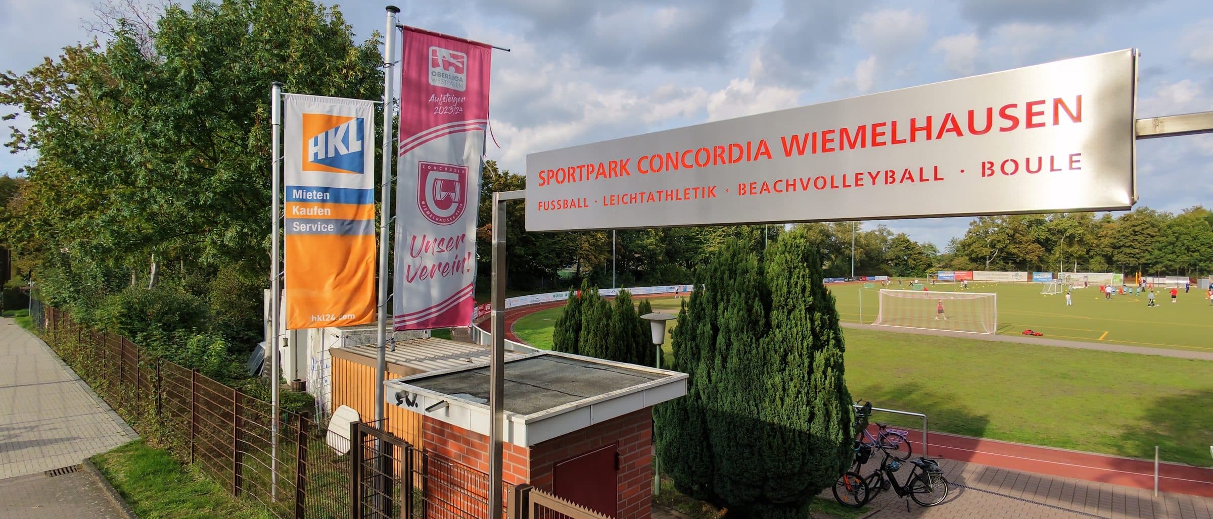 Eingang des Sportparks Concordia Wiemelhausen mit Schild, Fahnen und Blick auf den Kunstrasenplatz
